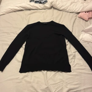 Svart långärmad tröja med vågig kant - Svart långärmad tröja från Aéryne med rund hals. Snygg och enkel design som är lätt att matcha med jeans eller kjol. Materialet känns mjukt och stretchigt, perfekt för dig som gillar stilrena plagg med en liten twist.