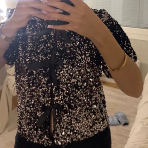 Paljett topp - Superfin svart blus täckt med glittrande paljetter och korta puffärmar. Framtill har den flera svarta rosetter som snygga detaljer. Från gina nypris är 400kr använd en gång 🩷