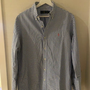 Blårutig skjorta från Ralph Lauren - Snygg blårutig skjorta från Ralph Lauren i storlek M. Skjortan har klassisk krage, vita knappar och det ikoniska broderade Polo-logot på bröstet i rött. Tillverkad i mjuk bomull med lång ärm och normal passform.