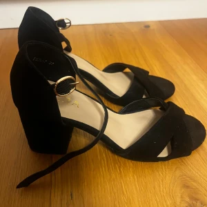 Svarta sandal klackar med klack från Cora - Snygga svarta sandaler från Cora i storlek 37 med bred klack och öppen tå. Skorna har en elegant remdesign och guldfärgat spänne vid ankeln. Materialet är mocka och sulan är platt framtill. Perfekt för dig som gillar stilrena och trendiga skor.