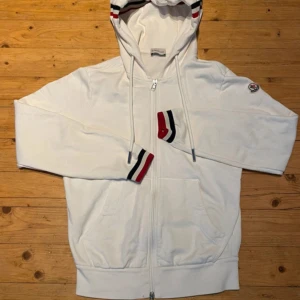 Moncler hoodie (ÄKTA) - Vit hoodie från Moncler med hel dragkedja, huva och fickor framtill. Ribbade muddar och huva med röda, blå och vita ränder. Klassisk Moncler-logga på ärmen. Perfekt för dig som gillar stilren streetwear med exklusiv känsla. Inga defekter, väldigt eftertraktad hoodie säljer nu den för ett bra pris. (Äkta) fraktar inom 24 timmar.