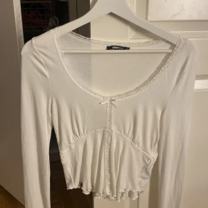 Vit långärmad blus från Gina Tricot - Supersöt vit blus från Gina Tricot i storlek S. Den har lång ärm, spetskant runt halsringningen, liten rosett framtill och volang vid nederkanten. Croppad passform som ger en trendig look. Perfekt att styla med jeans eller kjol. Ingen lapp kvar men aldrig använd.
