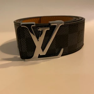 Louis Vuitton rutig skärp i skinn - Säljer ett Louis Vuitton bälte med klassiskt rutigt mönster i mörkgrå och svart. Bältet har en bred form och en stor, silverfärgad LV-spänne framtill. Insidan är beige och materialet är skinn. Perfekt för dig som vill ha en lyxig och stilren accessoar.