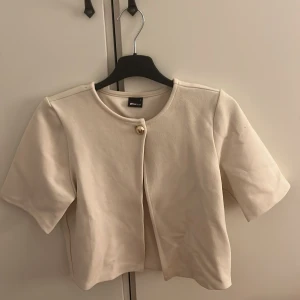 Beige croppad kavaj från Gina Tricot - Snygg beige croppad kavaj från Gina Tricot med korta ärmar och rund halsringning. Jackan har en guldfärgad knapp framtill och stilren design som ger en fräsch look. Perfekt att styla med jeans eller kjol för en trendig vibe.