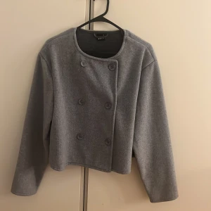 Grå kappa från Gina Tricot XS - Säljer en stilren grå kappa från Gina Tricot i storlek XS. Jackan har dubbelknäppning med sex knappar framtill, rund halsringning och långärmade ärmar. Perfekt för dig som gillar minimalistisk och clean look.
