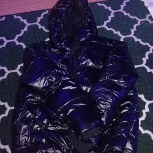 Svart pufferjacka med huva - Säljer en svart pufferjacka med glansig yta och huva. Jackan har lång ärm, dragkedja framtill och breda quiltade sektioner som ger en cool och modern look. Perfekt för kalla dagar när du vill hålla värmen och ändå se fresh ut. Använd 1 gång 