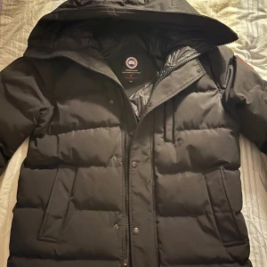 Svart dunjacka Canada Goose XL - Svart dunjacka från Canada Goose i storlek XL. Jackan har huva, dragkedja och tryckknappar framtill, samt två stora fickor. Klassisk patch på ärmen och logga på ryggen. Perfekt för kalla vinterdagar med sin tjocka, quiltade design.