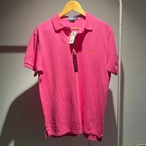 Rosa pikétröja från Ralph Lauren - Snygg rosa pikétröja från Ralph Lauren med klassisk krage och korta ärmar. Tröjan har den ikoniska broderade loggan i gult på bröstet och är gjord i mjuk bomull. Perfekt för dig som gillar färg och vill ha en stilren look.