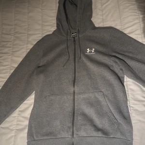 Under Armour Hoodie grå  -  Mörkgrå hoodie från Under Armour med dragkedja och broderad logga på bröstet. Tröjan har huva med snörning, två fickor fram och är fodrad med mjukt material. Perfekt för chill eller träning.