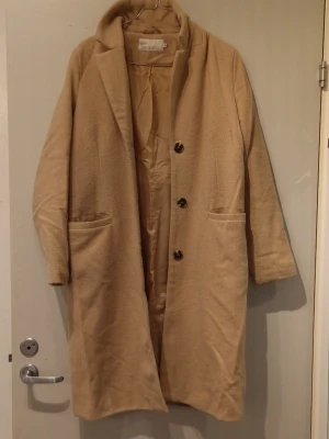 Beige kappa från NLY Trend, strl 34 - Snygg beige kappa från NLY Trend i storlek 34. Rak modell med krage, tre knappar framtill och två fickor. Materialet är 20% ull och 80% polyester, vilket gör den både mjuk och varm. Perfekt för höst och vinter när du vill ha en clean och stilren look. Den är noppig under ärmarna men annars i bra skick.