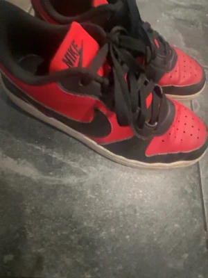 Nike Dunk sneakers röd och svart - Säljer ett par Nike Dunk sneakers i rött och svart med klassisk swoosh på sidan. Skorna har snörning, rund tå och platt sula. Ovandelen är i skinn och har perforerade detaljer på tån för extra ventilation. Perfekt för dig som gillar streetwear och sportig stil.