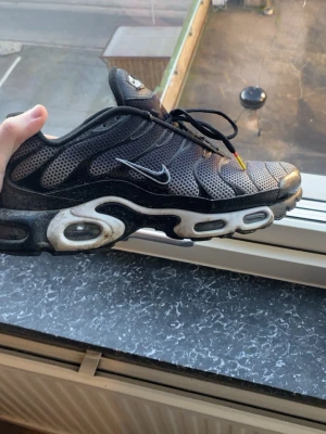 Nike Air Max Plus Tn sneakers svart - Nike Air Max Plus Tn sneakers i svart och grå mesh med silvriga detaljer och synlig Air Max-sula. Snyggt TN-märke på hälen och klassisk Swoosh på sidan. Skorna har snörning och en platt sula med rund tå. Perfekt för dig som gillar streetwear och sportig stil.