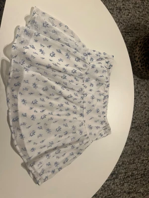 Vit blommig kortkjol från Gina Tricot - Supersöt vit kortkjol från Gina Tricot med blått blommigt mönster. Kjolen har en luftig och volangig design med resår i midjan för skön passform. Perfekt för dig som gillar romantisk och fräsch stil. Materialet känns lätt och somrigt.