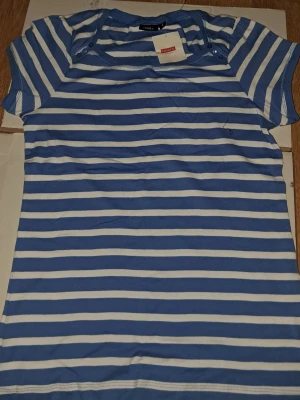 Blåvit randig t-shirt från Lindex med prislapp - Snygg blå och vit randig t-shirt från Lindex i storlek L. Modellen har korta ärmar och rund hals med tre små knappar på axeln som cool detalj. Materialet är mjuk bomull som känns skönt mot huden. Perfekt för dig som gillar klassisk stil med en twist.