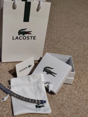 Lacoste silverfärgad länkarmband - Snyggt länkarmband från Lacoste i silverfärgad metall med detaljerad struktur på länkarna. Armbandet kommer med original ask och vit tygpåse med Lacoste-logga. Perfekt accessoar för dig som gillar stilrena och moderna smycken. Fortfarande i plastförpackning, ett till ett , fraktar snabbt:)