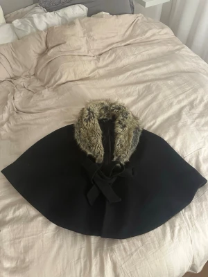 Svart kappa med fuskpäls och knyt - Säljer en svart kappa från H&M med bred krage i fuskpäls och snyggt knytband framtill. Kappan har en elegant och svepande form som ger en cool vibe, perfekt för dig som vill sticka ut lite extra. Pälsen är i beige och bruna toner.