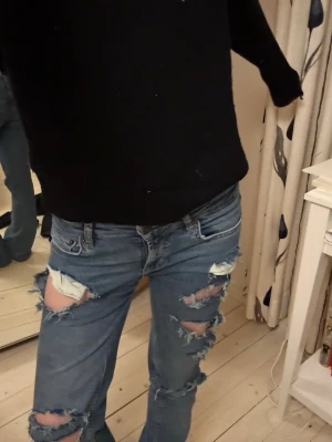 Blå slitna jeans med hål - Säljer ett par blå jeans med flera stora hål och slitningar på fram sidan. Jeansen har klassisk femficksdesign, knapp och dragkedja i midjan samt raka ben med lite bootcut-vibbar. Materialet är jeans och färgen är en mörkare blå tvätt. Byxerna har ja sytt ner själv 