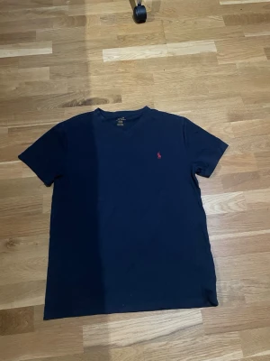 Mörkblå t-shirt från Polo Ralph Lauren - Snygg mörkblå t-shirt från Polo Ralph Lauren med klassisk röd broderad logga på bröstet. T-shirten har v-ringad hals och korta ärmar, tillverkad i mjuk bomull som känns skön mot huden. Perfekt för dig som gillar stilrena och ikoniska plagg.
