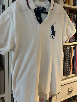 Vit pikétröja från Polo Ralph Lauren - Säljer en vit pikétröja från Polo Ralph Lauren i storlek M. Tröjan har klassisk krage med mörkblå knappslå och röda detaljer på kragen. På bröstet finns den ikoniska broderade polospelaren i marinblått. Perfekt för en clean och sportig look.
