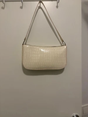 Beige axelväska med krokodilmönster - Snygg beige axelväska med krokodilmönster och glansig finish. Väskan har en avtagbar axelrem och en dragkedja upptill. Perfekt storlek för det viktigaste och ger en cool vibe till din outfit.