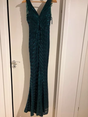 Glittrig långklänning med pärldetaljer - Säljer en mörkgrön långklänning med glittriga paljetter och snygga pärldetaljer vid axlarna. Klänningen har v-ringad fram och bak, är ärmlös och har ett mönster av glittriga ränder som ger en cool effekt. Perfekt för bal eller fest! Vi har också fixat den vid median 