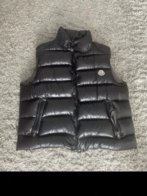 Svart dunväst från Moncler - Säljer en svart, glansig dunväst från Moncler med hög krage och två praktiska fickor med dragkedja. Västen har quiltad design och ikonisk Moncler-logga på bröstet. Perfekt för lager-på-lager och snygg till streetwear.Den är en storlek L/4 kan gå ner i pris om ni lägger ett bud👌knappt använd för den var för liten på mig 