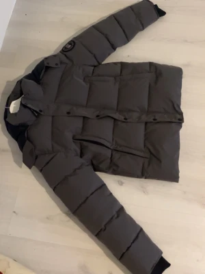 Mörkgrå pufferjacka från Canada Goose - Säljer en mörkgrå pufferjacka som är Canada Goose med klassisk patch på ärmen och hög krage. Jackan har långa ärmar, stora fickor och knappar framtill. Perfekt för kalla dagar och har en riktigt schysst oversized look.Fick som present har inte kommit till någon användning. Hör av dig vid frågor elller funderingar.