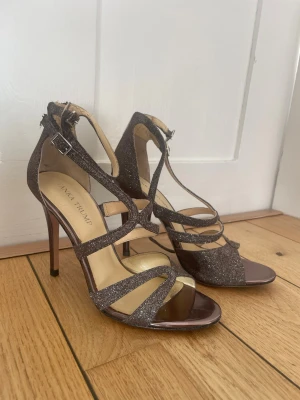 Glittriga bruna klackar  - Säljer ett par snygga sandaler från Ivanka Trump i mörkbrunt med glittrigt material. Skorna har hög klack, öppen tå och flera smala remmar med spänne runt ankeln. Perfekt för dig som vill sticka ut med lite extra glitter och elegans.
