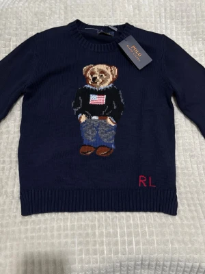 Mörkblå stickad tröja Polo Bear - Snygg mörkblå stickad tröja från Polo Ralph Lauren med ikonisk Polo Bear-motiv framtill. Rund hals och ribbade muddar. RL-broderi i rött nedtill. Perfekt för dig som gillar klassisk och lekfull stil med en twist. Helt oanvänd, pris kan diskuteras!