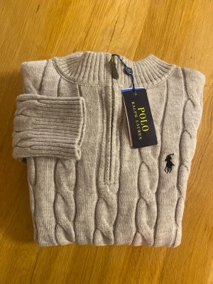 Beige stickad half zip tröja Polo Ralph Lauren - Säljer en grå stickad tröja från Polo Ralph Lauren i storlek M. Tröjan har ett klassiskt kabelstickat mönster, ribbade muddar och krage samt en half zip-dragkedja framtill. Ikoniska Polo-loggan broderad på bröstet. Perfekt för kyliga dagar.