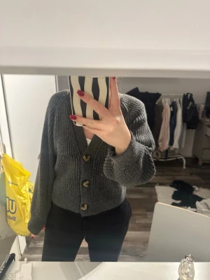 Grå stickad kofta med knappar - Säljer en mysig grå stickad kofta med tre stora träknappar framtill. Koftan har en v-ringad design och långa ärmar med breda muddar. Perfekt att slänga på sig när det är lite kyligt ute.