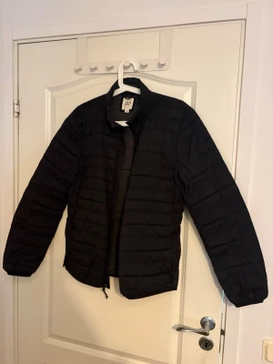 Svart pufferjacka från GAP - Svart pufferjacka från GAP med quiltad design och dragkedja framtill. Jackan har en enkel, stilren look med långärmade ärmar och lätt vaddering som gör den perfekt för kyliga dagar. Passar dig som gillar klassisk streetstil.