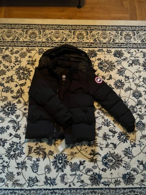 Svart pufferjacka från Canada Goose - Svart pufferjacka från Canada Goose i storlek M. Jackan har en stor huva, avtagbar pälsdetalj och klassisk logga på ärmen. Fylld med dun och har dragkedja samt knappar framtill. Perfekt för kalla dagar och har en clean, stilren look. Helt ny oanvänd. 