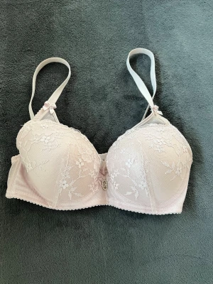 Ljusrosa bh - Säljer en ljusrosa bh  med spetsdetaljer och blommigt mönster. Bh:n har justerbara axelband, små rosetter och en dekorativ pärldetalj framtill. 