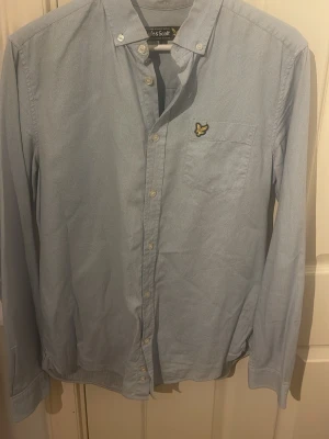 Ljusblå skjorta från Lyle & Scott - Snygg ljusblå skjorta från Lyle & Scott med diskret prickigt mönster och klassisk knappstängning. Bröstficka med gul broderad logga. Skjortan har lång ärm och button-down krage, perfekt för en chill och stilren look.