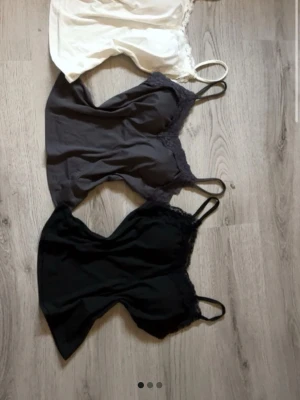 3 spetskantade linnetoppar i olika färger - Oanvända och nya toppar, stilrena linnetoppar med smala axelband och spetskant upptill. En topp är vit, en är grå och en är svart. Alla är ärmlösa och har en mjuk, slät yta med dekorativ spets längs halsringningen. Perfekt att matcha med jeans eller kjol.