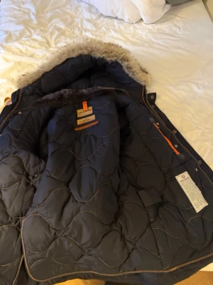 Parajumpers svart Vinter parkajacka med päls - Original Svart Vinter parkajacka köpt i Johnells från Parajumpers med snygg fuskpäls på huvan, orange detaljer och klassisk patch på ärmen. Jackan har dragkedja, knappar och flera fickor. Insidan är quiltad och jackan har en gul rem vid kragen. Perfekt för kalla dagar. 