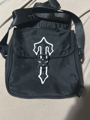 Svart axelväska från Trapstar - Trapstar axelväska i svart med justerbar axelrem och coolt emblem på locket. Väskan har meshdetaljer framtill, spänne med unik design och flera fack med dragkedja. Perfekt för att bära det viktigaste och matcha till streetwear.