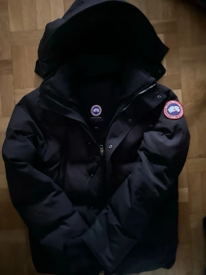 Svart pufferjacka från Canada Goose - Svart pufferjacka från Canada Goose med ikonisk patch på ärmen och stor huva. Jackan har dragkedja och tryckknappar framtill, samt flera praktiska fickor. Materialet är slitstark polyester och dunfyllning som håller dig varm under kalla dagar. Hör av dig vid intresse eller om du har nån fråga eller om du vill diskutera pris, råka ta bort alla täggs dom satt lösa 