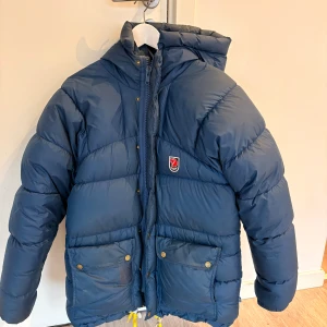  dunjacka från Fjällräven XXS - Säljer en blå dunjacka från Fjällräven med huva, stora fickor med tryckknappar och gul dragsko nedtill. Jackan har klassisk Fjällräven-logga på bröstet och är perfekt för kalla dagar. Fylld med dun och har dragkedja framtill. Obs! Se laglappar på bild. 