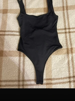 Svart ärmlös bodysuit - Säljer en svart bodysuit med breda axelband och djup urringning i ryggen. Modellen är tight och har en klassisk, stilren look som passar till många olika outfits. Materialet är mjukt och stretchigt, troligen polyester eller syntet. Perfekt för dig som gillar minimalistisk stil.