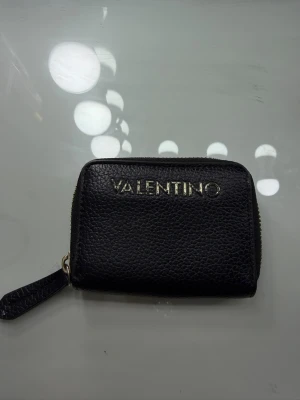 Svart plånbok från Valentino - Snygg svart plånbok från Valentino i skinn med guldfärgad logga på framsidan. Plånboken har dragkedja runtom och flera fack inuti för kort och sedlar. Perfekt storlek för att ha med sig överallt och stilren design som passar till allt.