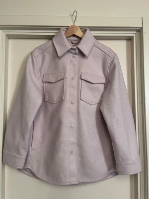 Ljuslila overshirt jacka - Stilren ljuslila overshirt jacka med stora bröstfickor och tryckknappar framtill. Jackan har klassisk krage, långa ärmar med knappdetalj och är gjord i ett mjukt ull-liknande material. Perfekt för lager-på-lager eller som vårjacka / över klänning på sommaren. XS men oversize som en M. Nyskick! Säljs på fler sidor, först till kvarn. 