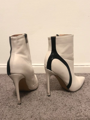 Vita stövletter med svart detalj - Säljer ett par vita stövletter med höga smala klackar och spetsig tå. Skorna har svarta, glansiga detaljer längs sidan och dragkedja på insidan. Materialet är slätt skinn och designen är riktigt edgy och modern.