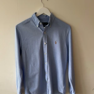 Blå oxfordskjorta från Ralph Lauren - Snygg ljusblå oxfordskjorta från Ralph Lauren med button-down krage. Skjortan är i storlek S och SP, den passar till alla tillfällen och alla årstider pågrund av sin lyxiga style och look. 