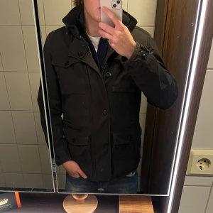 Svart parkajacka från Marc O'Polo - Svart parkajacka från Marc O'Polo i storlek M. Jackan har hög krage, flera fickor med lock och knappar samt dragkedja. Materialet är bomull och polyester, perfekt för kyliga dagar. Snygg och stilren design som funkar till jeans och hoodie.