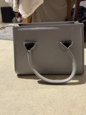 Grå handväska med dragkedja - Snygg grå handväska i skinnimitation med stilren, boxig form. Väskan har två korta handtag och en avtagbar axelrem med guldfärgade detaljer. Insidan har två fack med dragkedja för smidig organisering av dina grejer. Perfekt storlek för det viktigaste.