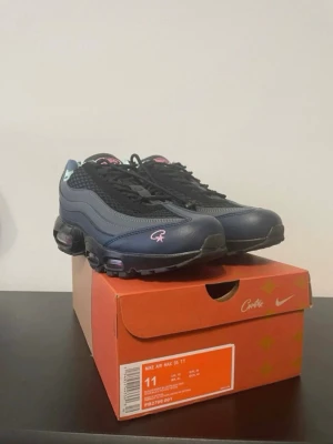 Nike Air Max 95 corteiz - Nike Air Max 95 corteiz sneakers i svart och mörkblå med rosa detaljer och logga. Skorna har snörning, platt sula och rund tå. Insidan har ett camouflagemönster i blå toner och rosa Nike Air-tryck. Luftkuddar i sulan för extra komfort. Perfekt för dig som gillar streetwear.
