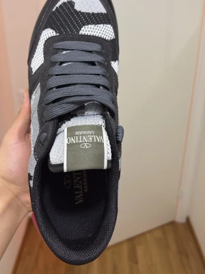 Valentino Garavani Rockrunner sneakers - Säljer ett par Valentino Garavani Rockrunner sneakers med svart, grå och vit camouflage-mönster. Skorna har snörning, platt sula med nitar på sidan och en röd detalj på hälen. Materialmix av mocka och syntet, rund tå och cool streetstil.