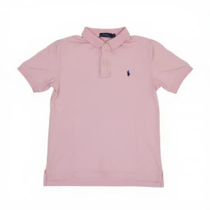 Rosa piké från Polo Ralph Lauren - Säljer en ljusrosa pikétröja från Polo Ralph Lauren med klassisk krage och korta ärmar. Tröjan har knappar framtill och den ikoniska broderade loggan i mörkblått på bröstet. Perfekt för en chill och stilren look. (Storlek junior M (10-12)